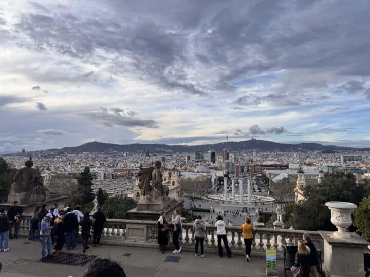 People stand on a terrace overlooking a family friendly Barcelona cityscape, with statues in the foreground, distant mountains, and a cloudy sky. Below are tall buildings and columns, blending historic charm with modern attractions perfect for all ages.