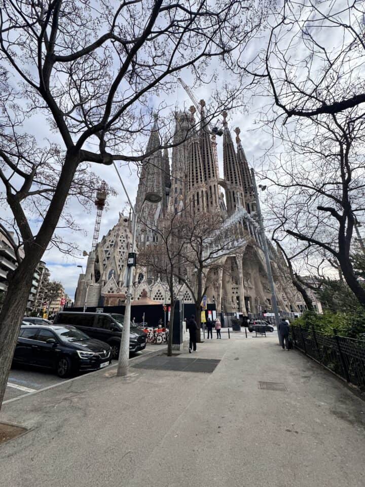 A wide view of the Sagrada Família basilica in Barcelona, showing its ornate spires and ongoing construction—an iconic stop for a Barcelona family vacation—amid bare trees, parked cars, and people walking along the adjacent sidewalk.