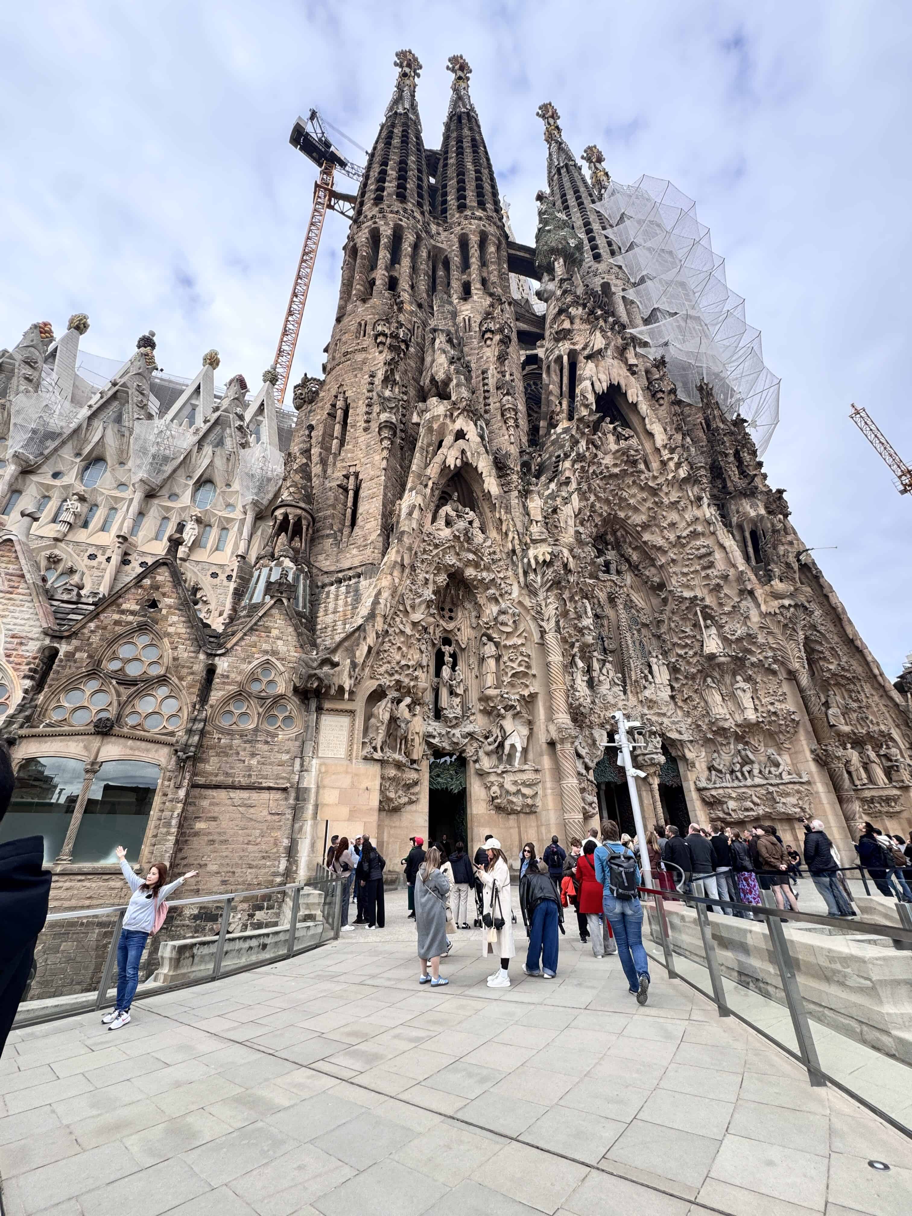 Tourists gather at the entrance of the Sagrada Familia in Barcelona, Spain. The ornate stone façade and towers make it a highlight for family travel Barcelona, with ongoing construction visible under mostly cloudy skies.