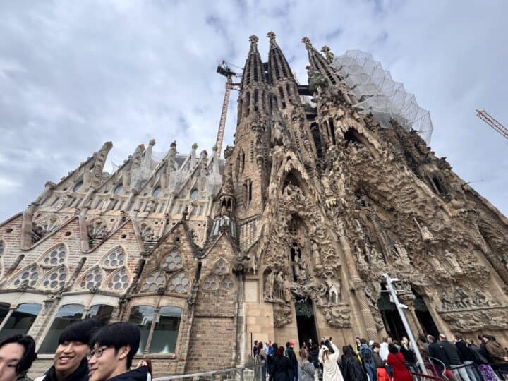 A wide-angle view of the Sagrada Familia basilica in Barcelona, showing its intricate facades, tall spires, a construction crane, and groups of people—perfect for a Family-Friendly Barcelona outing—gathered near the entrance under a cloudy sky.