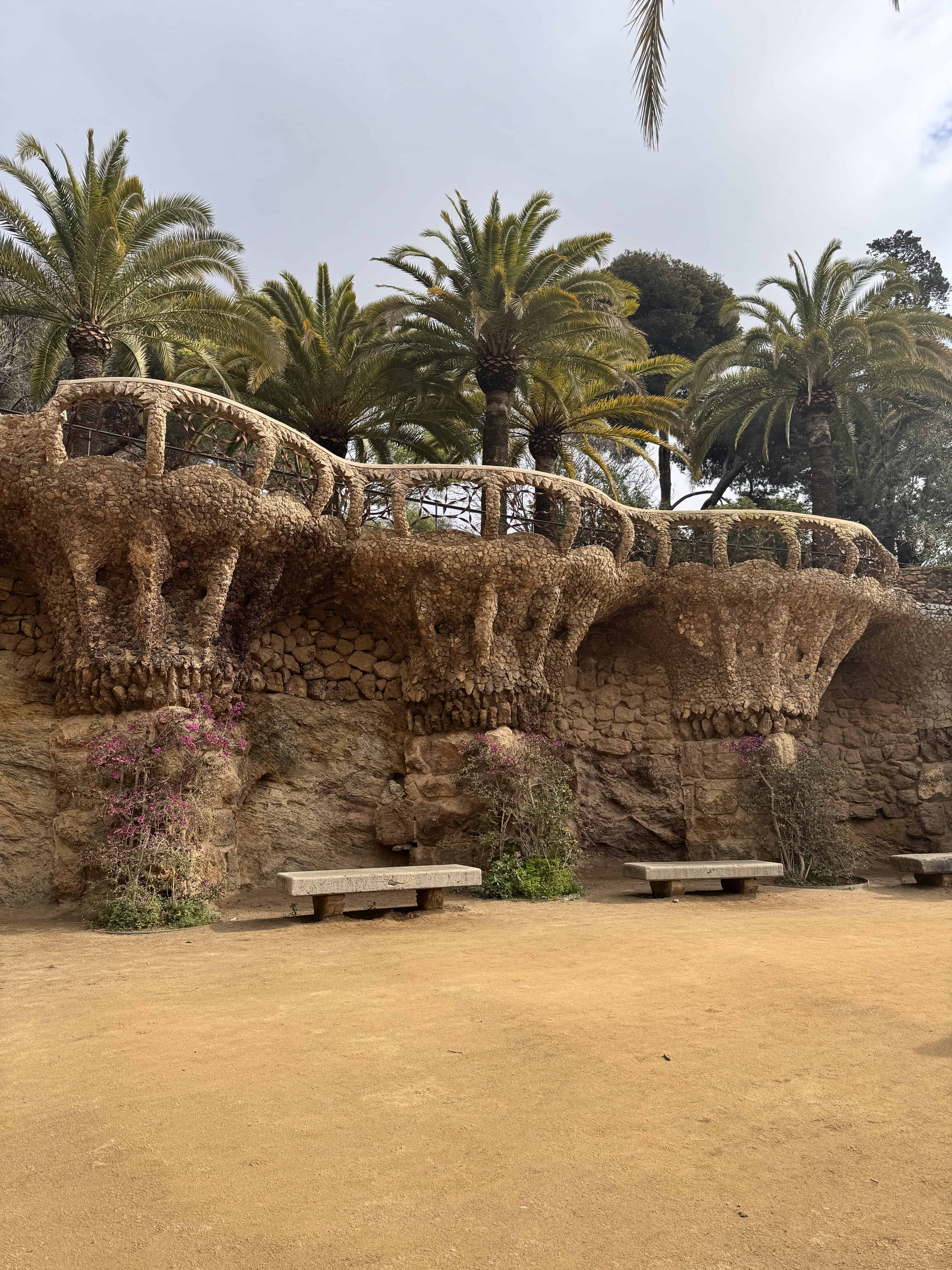 A stone structure with unique, curved supports stands in front of tall palm trees. Perfect for a Barcelona family vacation, three stone benches sit by the base while flowering plants grow on sandy ground under a cloudy sky.