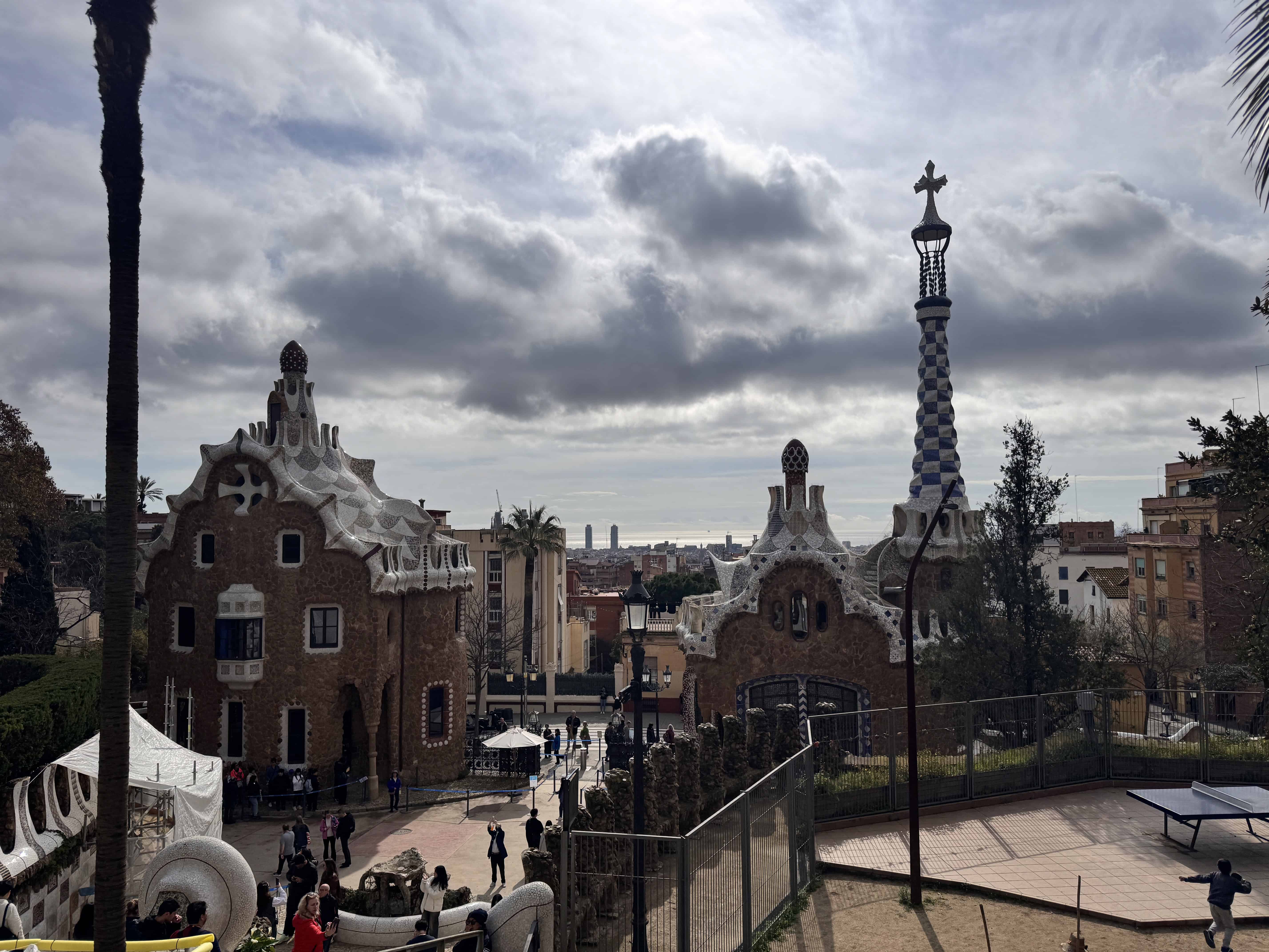 Enjoy a family-friendly Barcelona adventure at Park Güell, where kids can marvel at the wavy-roofed buildings and tall column topped with a cross, while families stroll and gather on this cloudy day.