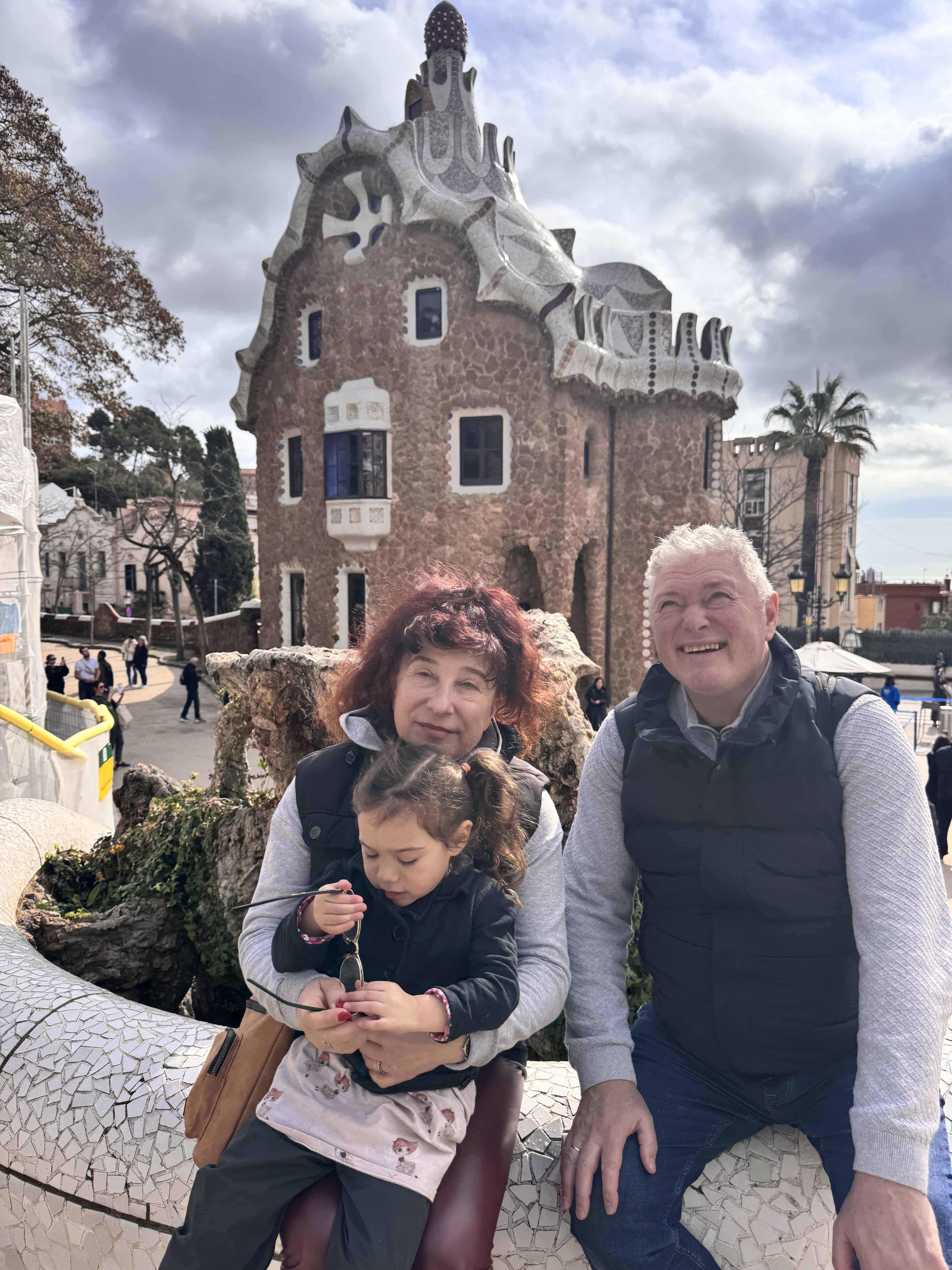 Three people sit on a mosaic bench in front of a distinctive building in Park Güell, enjoying a family-friendly Barcelona moment. An older woman holds a small child while an older man sits beside them, all dressed casually under a partly cloudy sky.