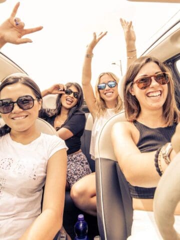 Four women wearing sunglasses smile and pose inside a car with the roof open. One is driving, while the others make celebratory gestures. It appears to be a sunny day, and the car is parked in a rural area.