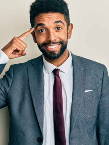 A man in a suit and tie stands in front of a light-colored wall, smiling and pointing to his temple with his right index finger.