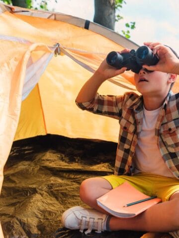 A young boy sits cross-legged at the entrance of a yellow tent in a grassy, wooded area, looking through binoculars. A notebook and pencil rest on his knee.