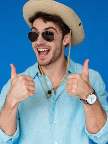 A man wearing a straw hat, sunglasses, and a light blue shirt stands in front of a blue background, smiling broadly and giving two thumbs up.