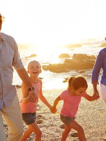 Two adults and two children walk hand-in-hand along a beach at sunset. The adults wear sunglasses and summer clothing, and everyone appears to be smiling. The ocean and rocks are visible in the background.