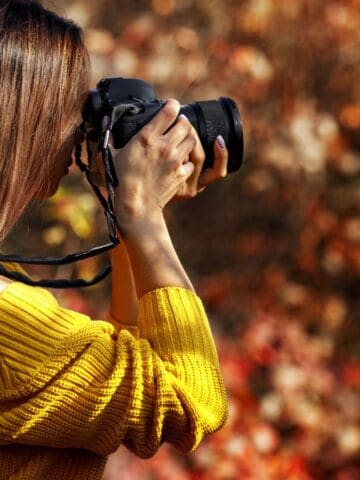 A person with long brown hair wearing a yellow sweater is holding a camera up to their face, taking a photograph outdoors with a blurred background of red and orange foliage.