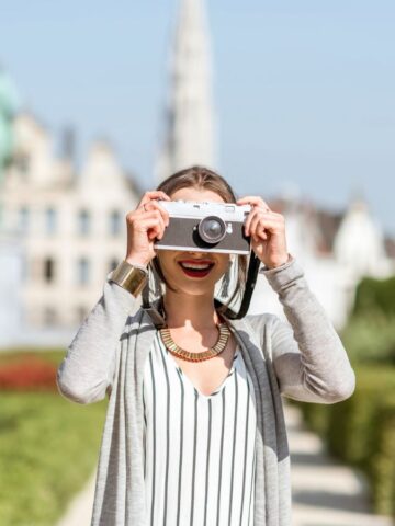 A woman standing outdoors in a garden holds a camera up to her face, taking a photo. Buildings and a statue are visible in the blurred background on a sunny day.