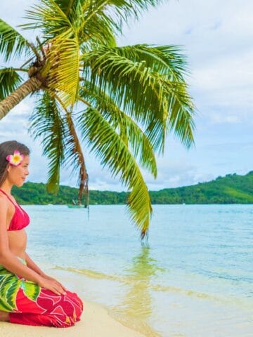A woman in a red bikini top and floral wrap sits on a sandy beach near a leaning palm tree, facing the calm ocean. Lush green hills and trees are visible across the water under a partly cloudy sky.