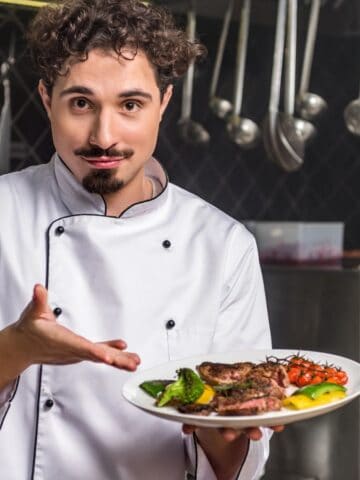 A chef in a white uniform stands in a commercial kitchen, holding a plate of cooked meat with vegetables and gesturing toward the dish, with various kitchen tools hanging in the background.