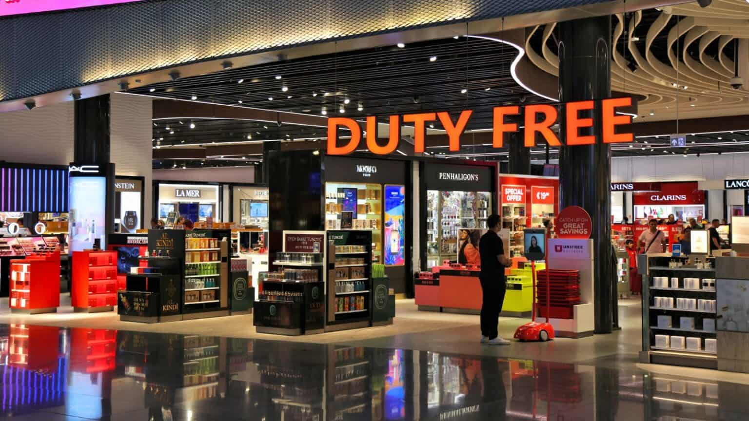 A brightly lit airport duty-free store displays various products such as perfumes, cosmetics, and liquor. Shoppers browse the shelves, and a large orange 