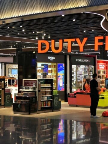 A brightly lit airport duty-free store displays various products such as perfumes, cosmetics, and liquor. Shoppers browse the shelves, and a large orange "DUTY FREE" sign hangs above the entrance.