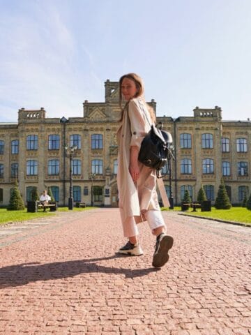 A woman wearing light-colored clothes and a backpack walks along a red brick path toward the camera, with a large historic building and green lawn in the background. The sky is clear and sunny.