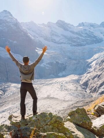 A person stands on a rocky ledge with arms raised, facing a snowy mountain range and glacier under a clear sky. Autumn-colored trees are visible on the right side of the foreground.