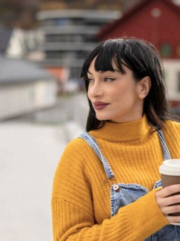 A woman wearing a yellow turtleneck sweater and denim overalls stands on a bridge holding a disposable coffee cup, looking to the side with buildings in the background.
