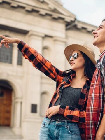 Two people in casual clothes and sunglasses, one with a backpack, stand in front of a large historic stone building. The woman points ahead while the man looks in the same direction.