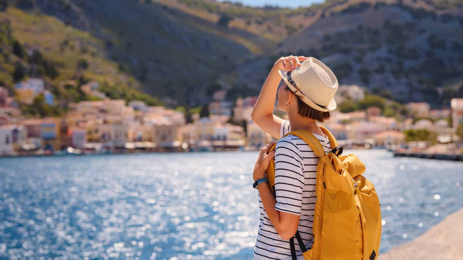 A person wearing a striped shirt, yellow backpack, and hat stands by the water, looking toward a coastal town with colorful buildings at the base of a mountain.