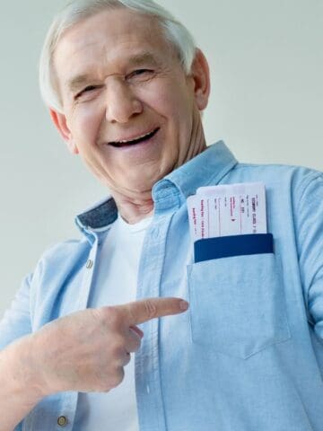 An older man smiles while pointing to airline tickets in his shirt pocket and giving a thumbs-up gesture. He is wearing a light blue button-up shirt and standing against a plain, light background.