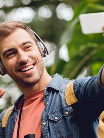 A young man wearing headphones and a backpack smiles while taking a selfie outdoors. He is making an "OK" gesture with his hand and stands in front of green foliage.