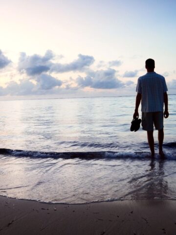 A man stands at the edge of the ocean during sunset or sunrise, holding his shoes and looking at the water. The beach is mostly empty, and the sky is partly cloudy with soft light.