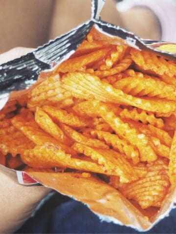 A hand holds an open bag of crinkle-cut, orange seasoned potato chips—perfect airport snacks. Blue denim and part of a leg and shoe are visible in the background.