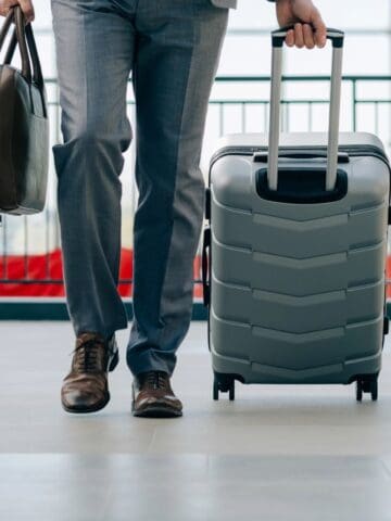 A person wearing a suit walks indoors carrying a suitcase in one hand and a briefcase in the other, perhaps on business in countries where life is better than the Canadian standard. The background shows large windows and railings.