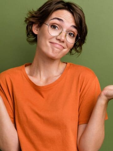 A person with short brown hair and glasses wearing an orange shirt stands against a green background, raising both hands with palms up and shoulders slightly lifted, as if expressing uncertainty.