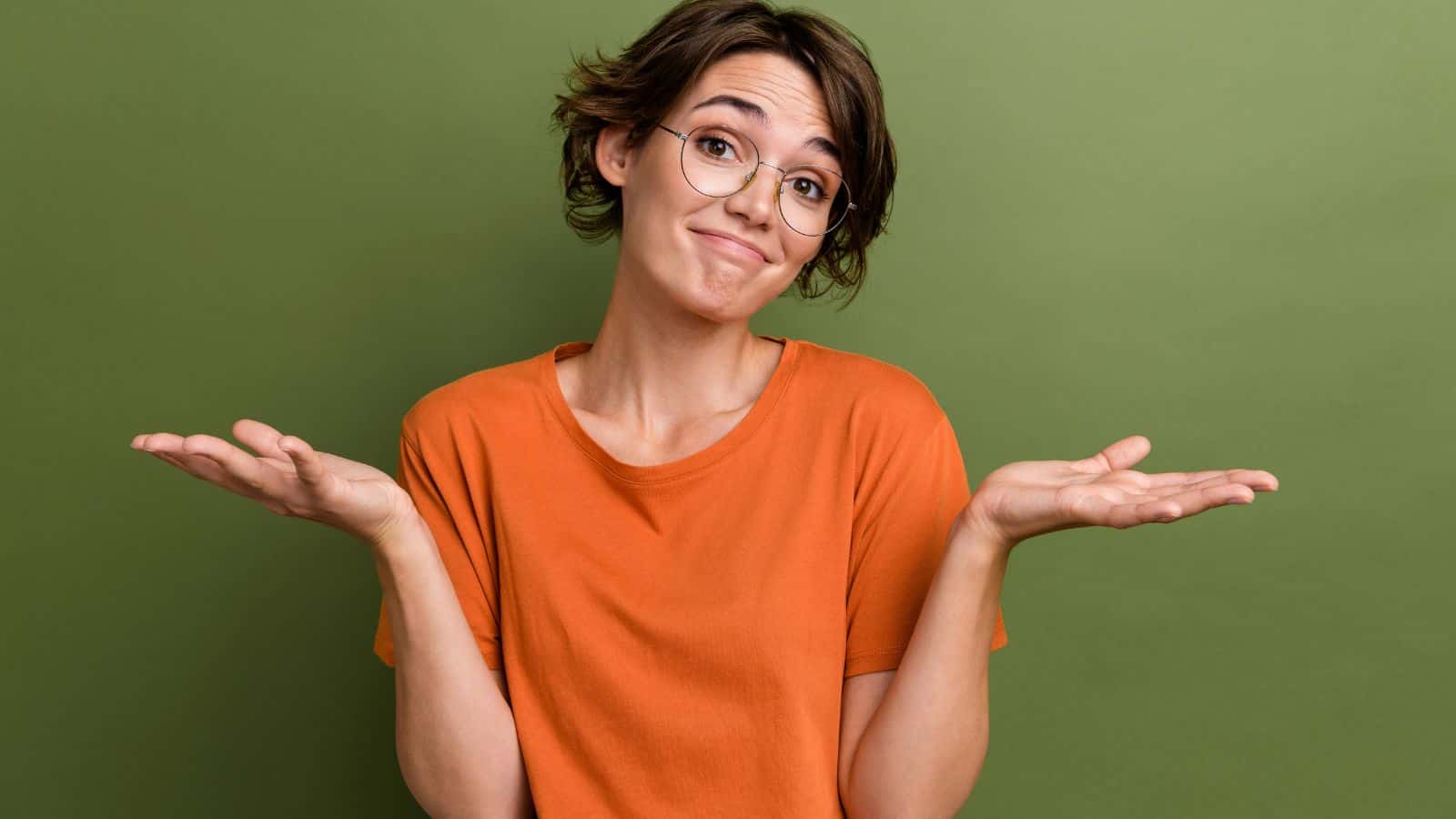 A person with short brown hair and glasses wearing an orange shirt stands against a green background, raising both hands with palms up and shoulders slightly lifted, as if expressing uncertainty.