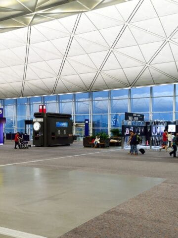 People walk with luggage inside a spacious, modern airport terminal with high ceilings, large windows, and bright lighting. Boarding gates and information displays are visible in the background.