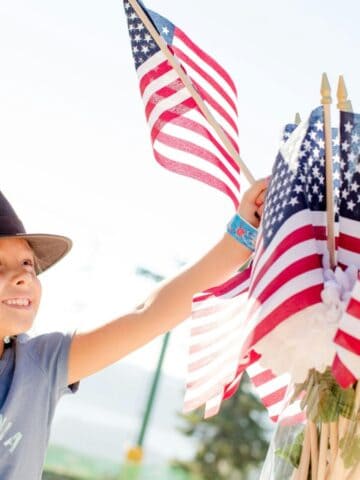 A young child wearing a black hat and blue "California" t-shirt smiles and reaches toward a bouquet of American flags outdoors on a sunny day, capturing the joy of local 4th of July celebrations.