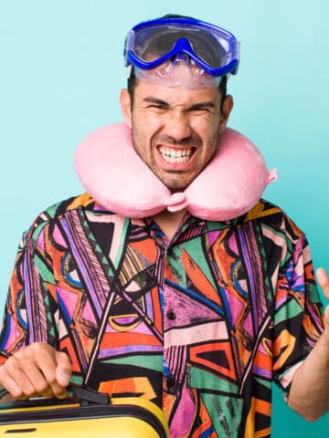 A man wearing a colorful shirt, a pink neck pillow, and blue swim goggles holds a yellow suitcase. He has an annoyed expression and his other hand is raised as if frustrated, standing in front of a blue background.