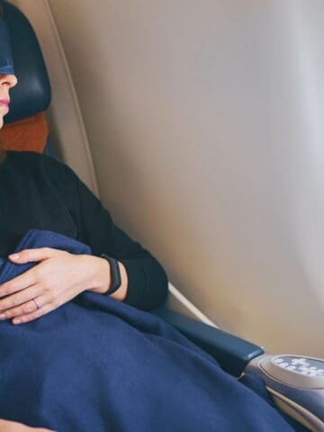 A person wearing an eye mask is sitting in an airplane seat, covered with a dark blue blanket, resting with their hands on their lap near a window.