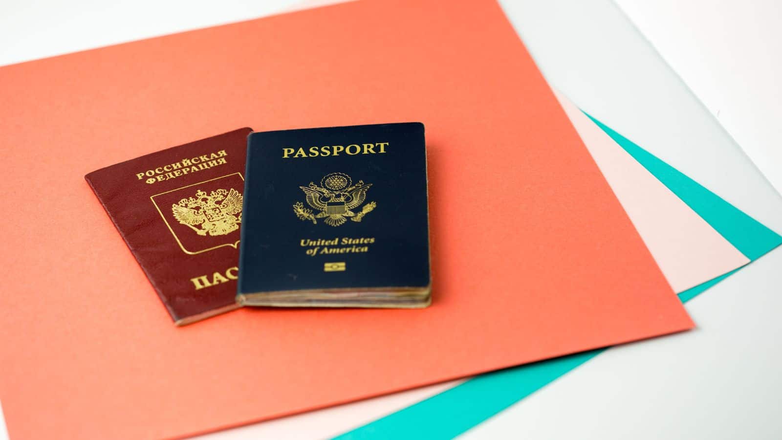 A Russian Federation passport and a United States of America passport are placed side by side on top of red, pink, and teal colored sheets of paper.
