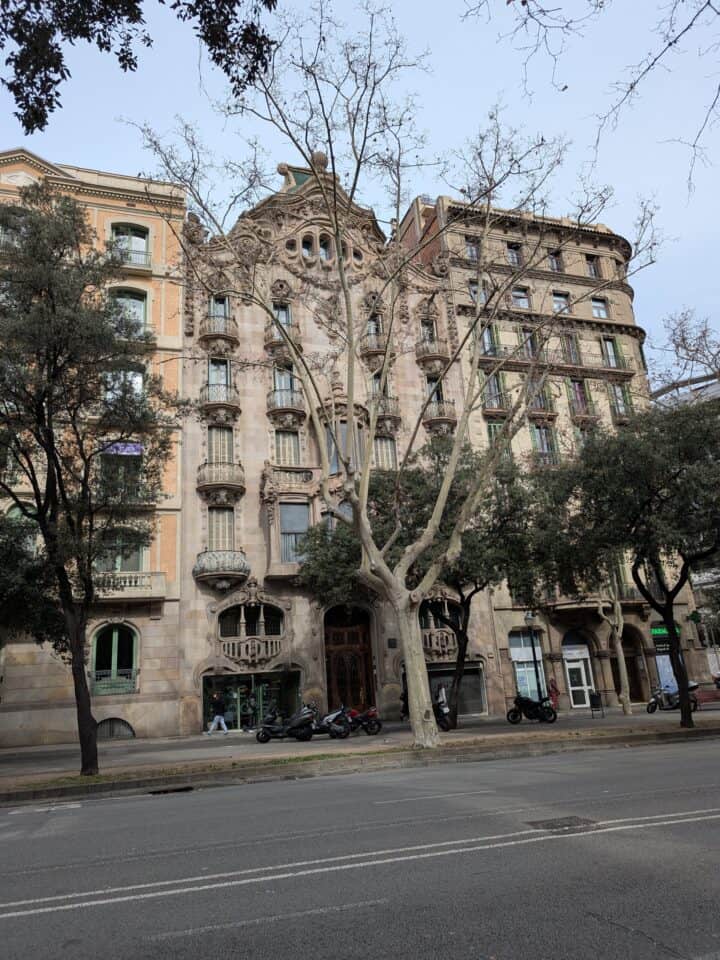 A stone building with ornate balconies and arched windows stands behind leafless trees. Parked scooters line the sidewalk of this quiet street, perfect for a family-friendly Barcelona stroll.