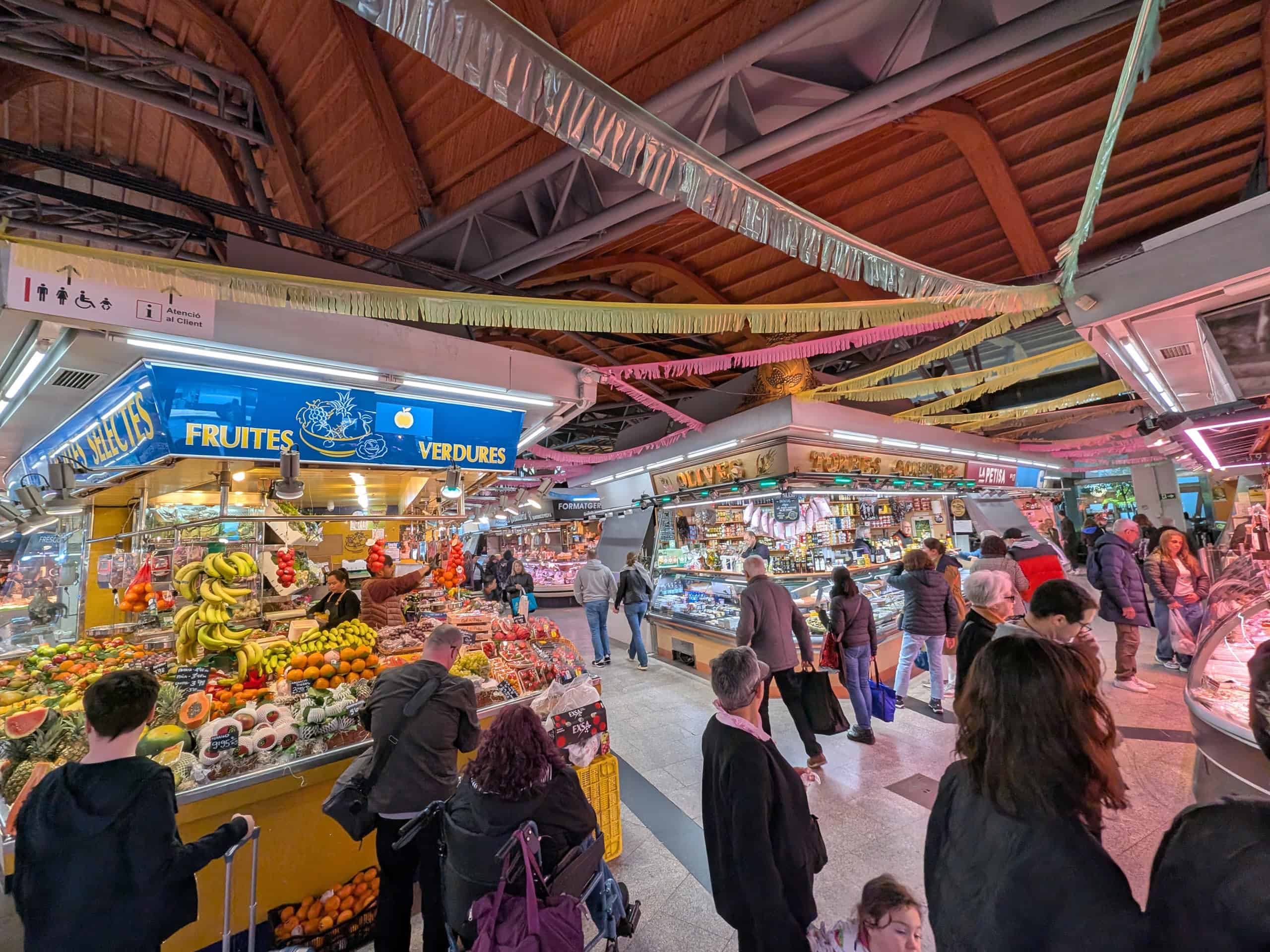 Indoor market scene in family-friendly Barcelona with stalls selling fruits, vegetables, and groceries. Shoppers, including families on a Barcelona family vacation, browse busy aisles under curved wooden beams and multilingual signs.