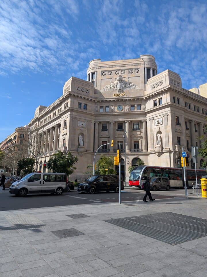 A large stone building with ornate architectural details and a "BBVA" sign on its facade, located at a bustling street intersection—a classic scene for a family-friendly Barcelona adventure, with cars, taxis, pedestrians, and a red city bus nearby.