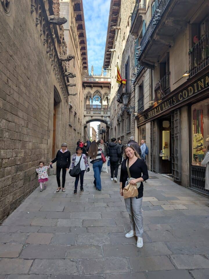 A woman stands and smiles on a narrow stone street lined with historic buildings in family-friendly Barcelona. Several people walk along the street, and a bridge connects two buildings in the background. A Spanish flag hangs from a balcony.