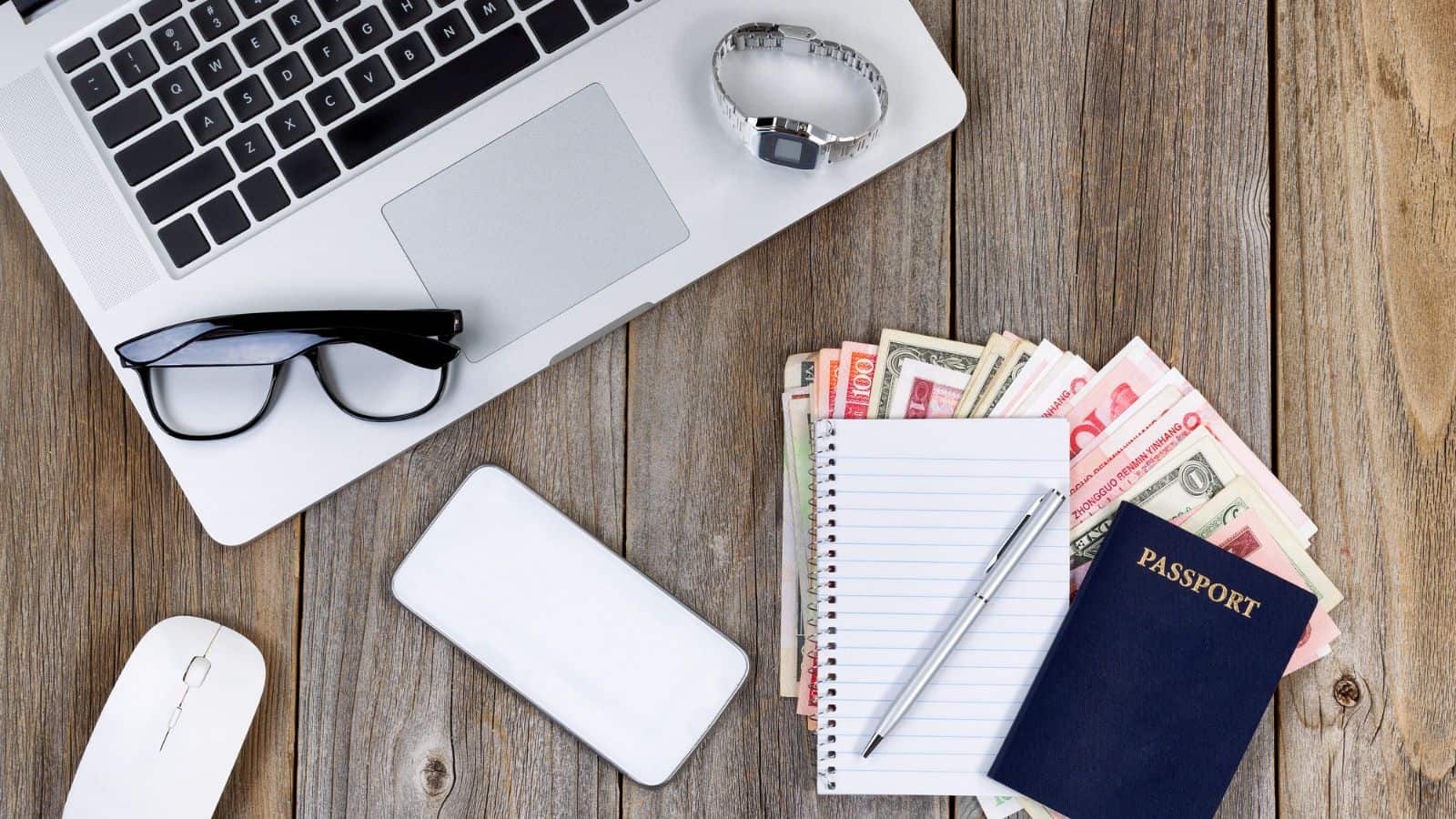 A laptop, glasses, smartphone, mouse, wristwatch, notepad with pen, various paper currencies, and a passport are arranged on a wooden surface.