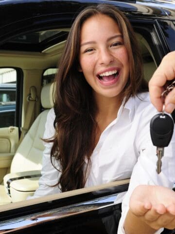 A person sitting in the driver’s seat of a black rental car smiles as another hand passes them a key through the window. The beige interior adds comfort, making this a top choice for smart travelers. Other cars are visible in the background.