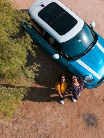 Aerial view of two people standing beside a blue car with a white roof, parked on a sandy surface near green foliage. Both look up toward the camera.