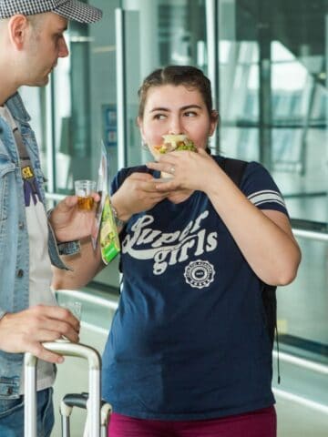 A man in a denim jacket and cap stands with a suitcase, while a woman beside him eats a sandwich. Another person, partially visible, stands nearby. They appear to be in an airport or transit area.