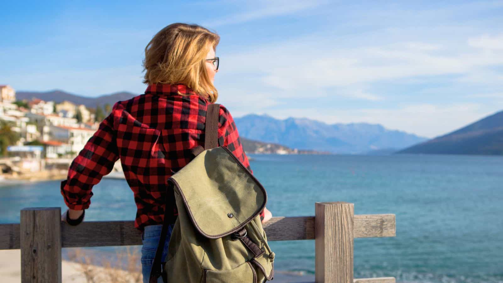 A person wearing a red and black plaid shirt and a green backpack leans on a wooden fence, looking out over a coastal landscape with blue water, mountains, and buildings in the distance.