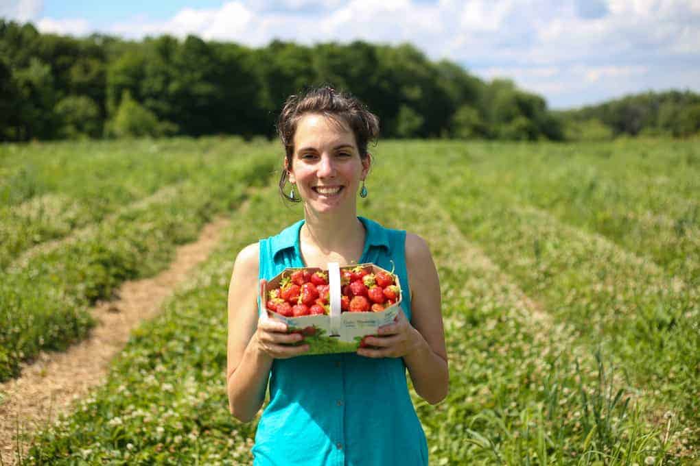 A person stands in a strawberry field holding a basket of freshly picked strawberries and smiling at the camera. Trees and a partly cloudy sky are visible in the background.