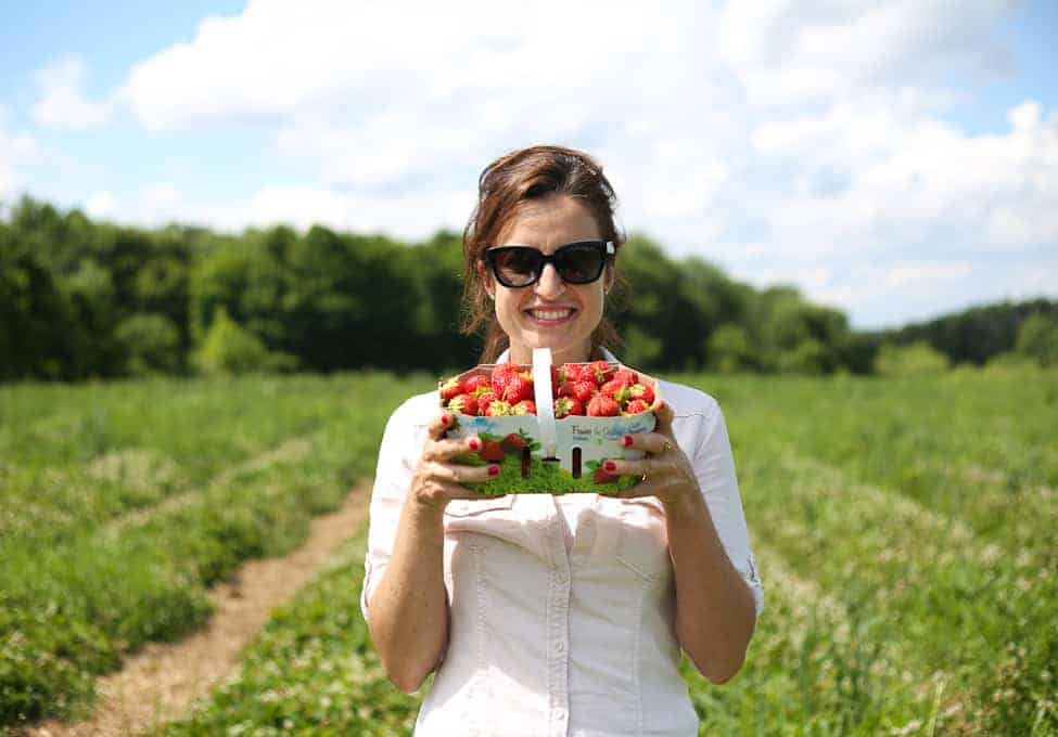 A woman wearing sunglasses and a white shirt stands in a field, holding two baskets filled with strawberries. The sky is partly cloudy and trees line the background.