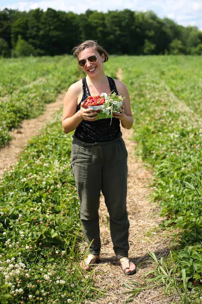 A person stands in a green field holding a bowl filled with strawberries and greens. They are wearing sunglasses, a black sleeveless top, and gray pants, and smiling at the camera. The sky is partly cloudy.