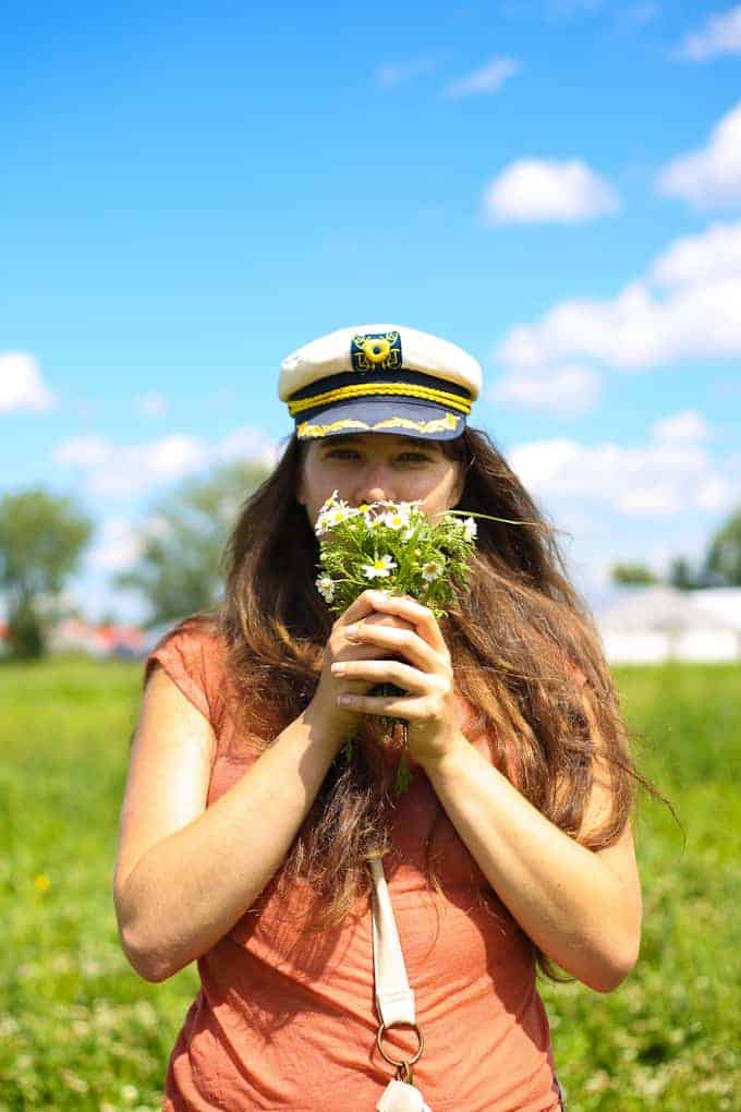 A person with long brown hair, wearing a white and blue captain's hat and an orange shirt, stands in a green field holding a small bouquet of wildflowers up to their face. The sky is blue with scattered clouds.