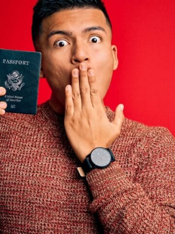 A person wearing a brown sweater and watch holds a United States passport in one hand while covering their mouth with the other, possibly surprised by new visa insights, standing in front of a solid red background.