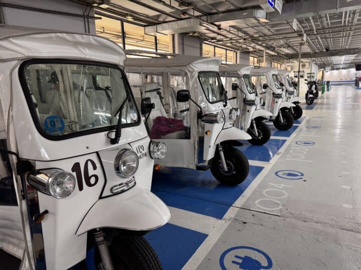 A row of white electric tuk-tuks is parked in numbered spaces inside a covered parking area. The ground and signs indicate that the parking is designated for electric vehicles—a convenient option for exploring family-friendly Barcelona.